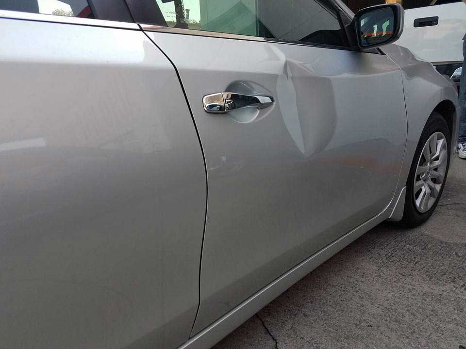 A side view of a silver sedan with a prominent dent on the front passenger side door panel near the door handle.