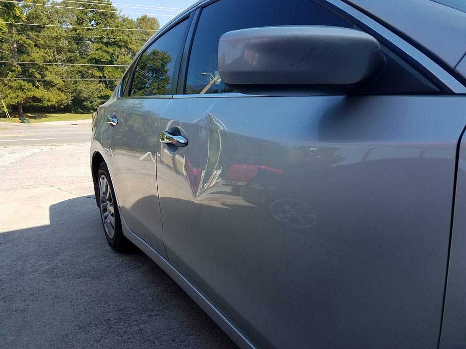 A silver sedan parked outdoors, showing a noticeable dent in the passenger side door below the side mirror.