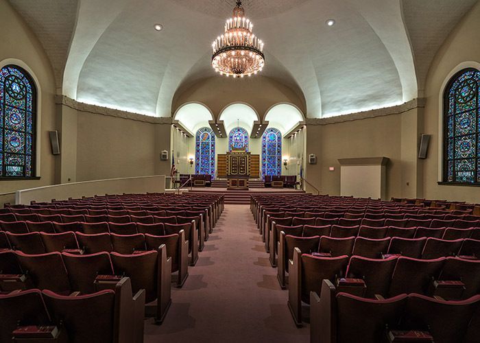 Interior of a synagogue with rows of seats facing a raised platform, a chandelier hangs from the ceiling.