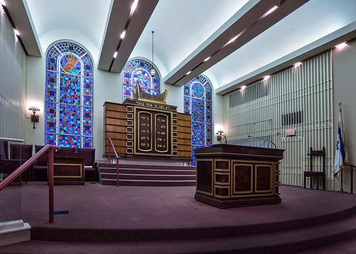 Interior of a synagogue sanctuary with stained glass windows, a Torah ark, and a bimah on a red carpet.