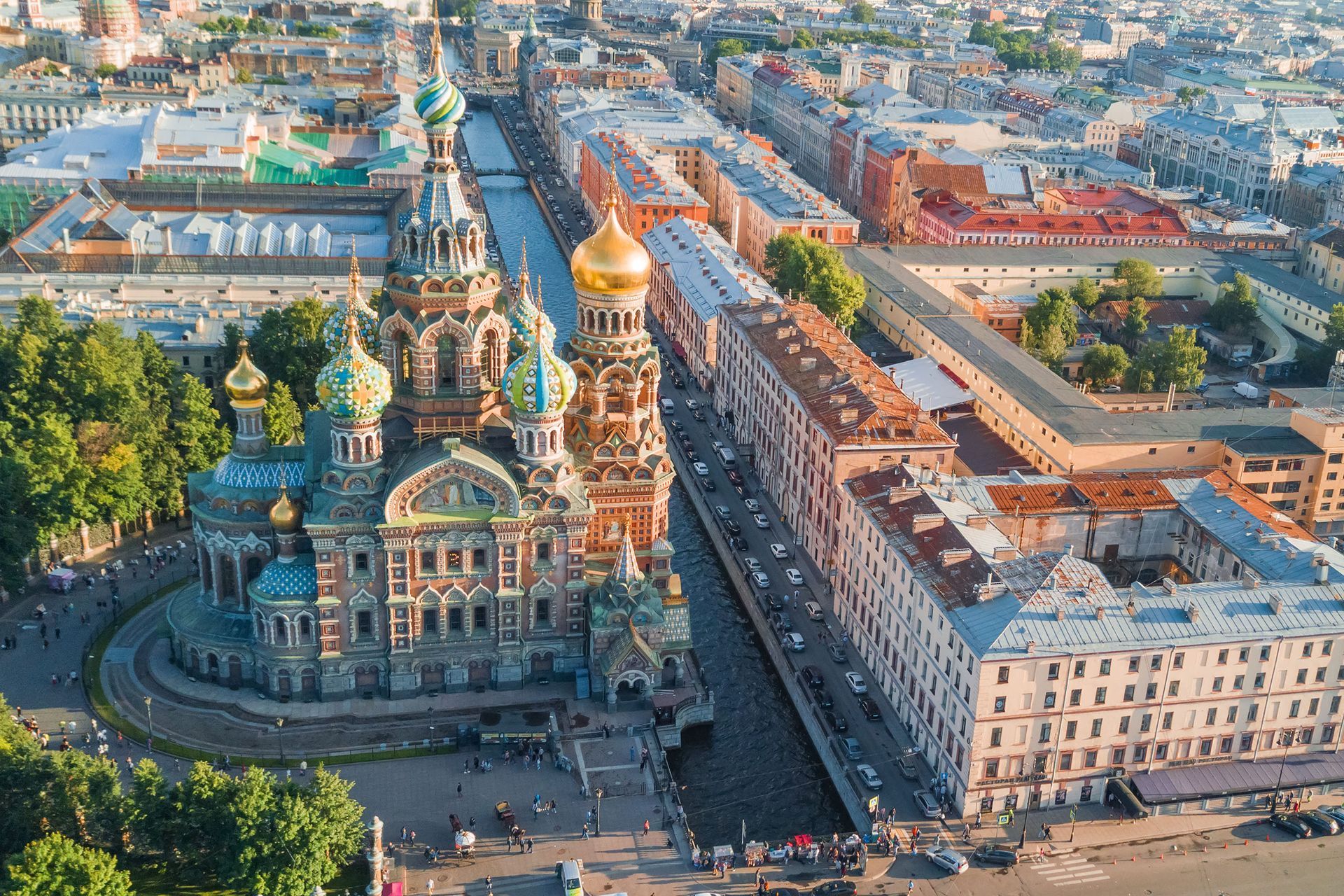 Church of the Savior on Spilled Blood, St. Petersburg, Russia, alongside a canal and buildings, with people, trees, and blue sky.
