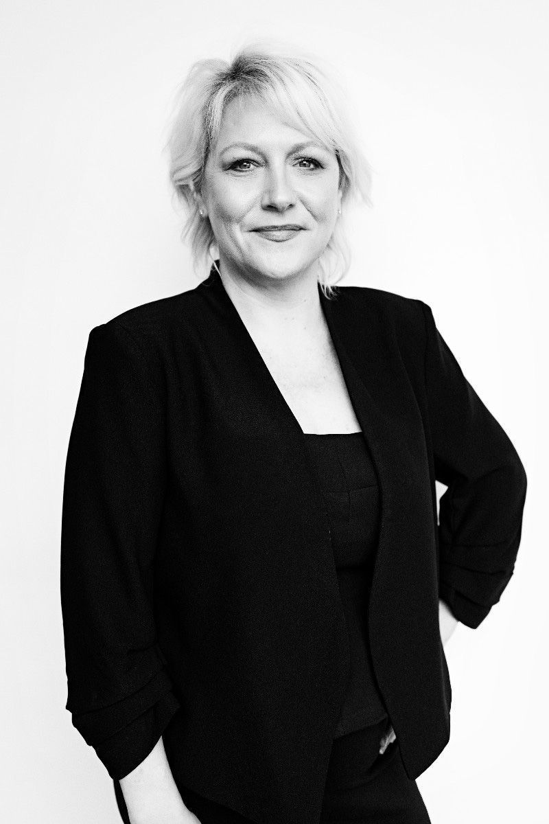 Woman with short blonde hair in a black blazer, poses with one hand on hip, smiling.