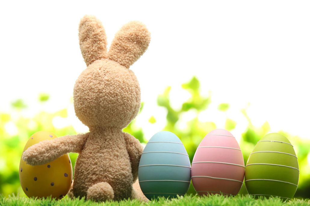 A stuffed bunny sits in green grass next to four colorful, decorated Easter eggs against a bright, blurred background.
