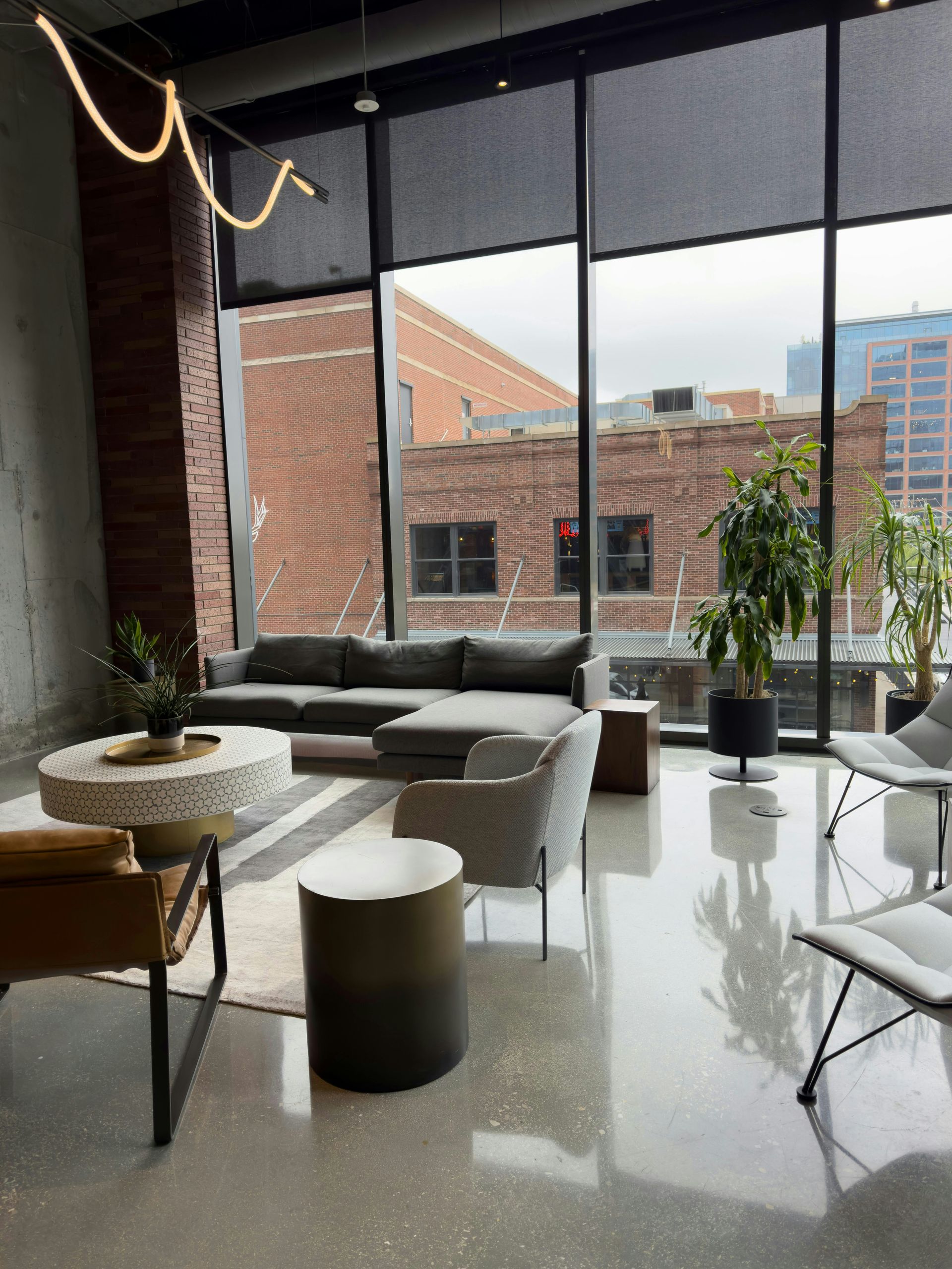 Modern lounge with gray sofas, white tables, large windows, and city brick buildings outside.