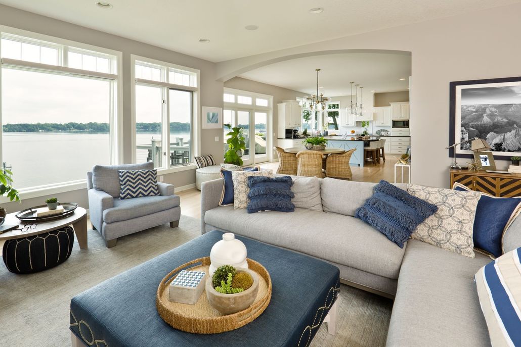 Bright living room with a curved gray sofa, blue accents, lake view windows, and an open kitchen beyond.