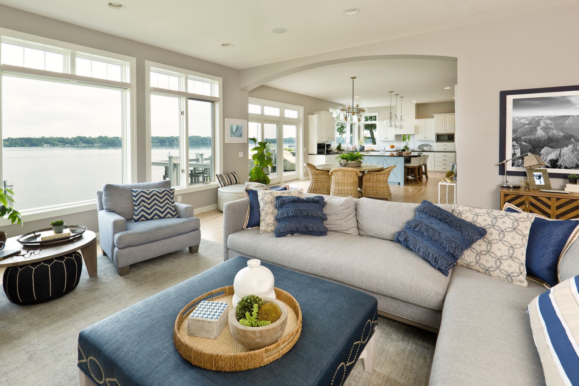 Bright living room with a curved gray sofa, blue accents, lake view windows, and an open kitchen beyond.