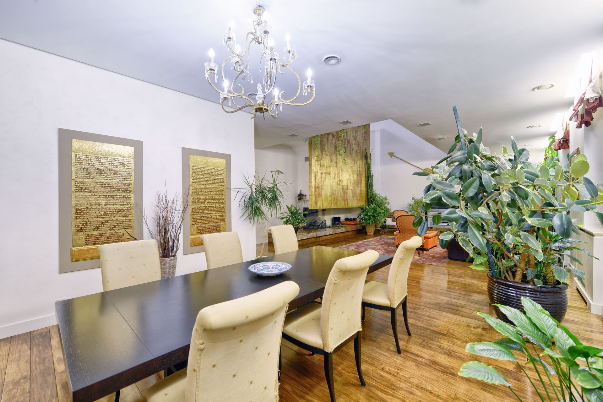 A dining room with a long table, cream-colored chairs, a crystal chandelier, gold wall art, and large indoor plants.