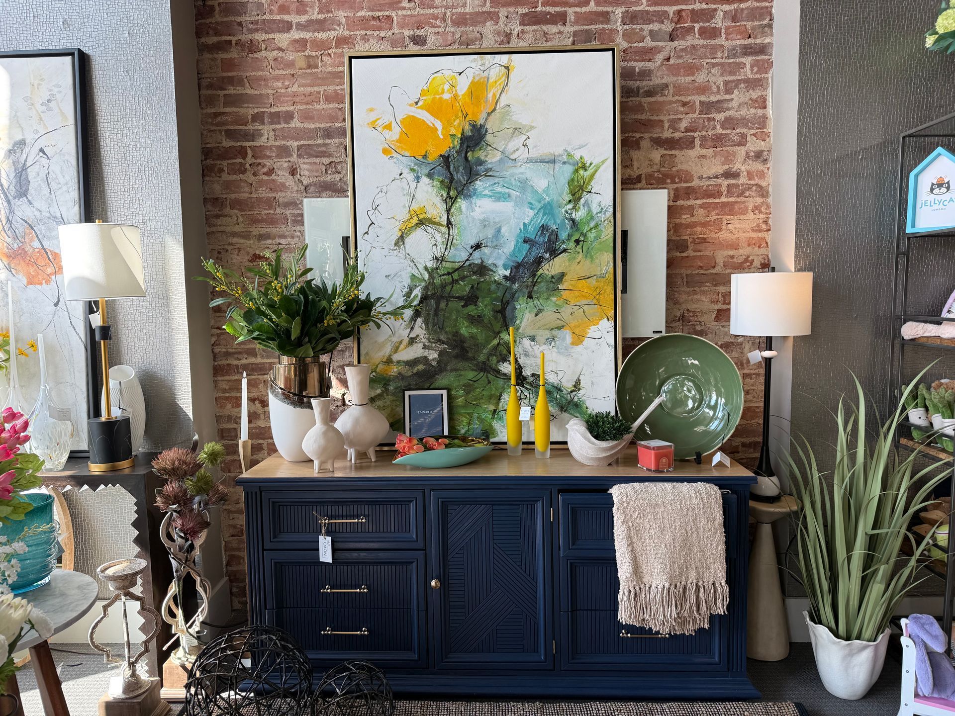 Stylish living room vignette with navy cabinet, abstract art, plants, and table lamps against a brick wall