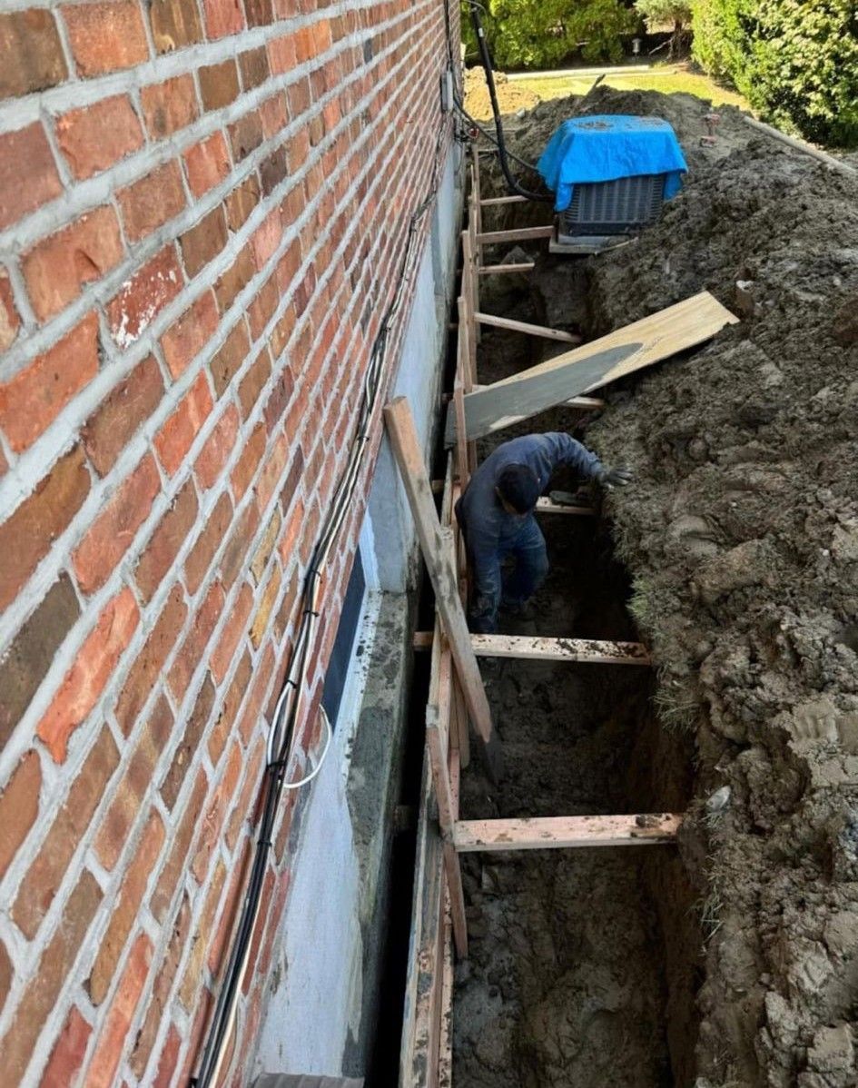 Narrow trench beside brick house with wooden braces, a worker inside, and a blue tarp-covered pile at the end