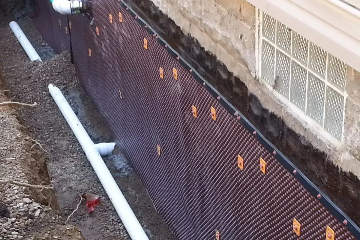 Black drainage membrane along a foundation wall beside white PVC pipes and dirt trench