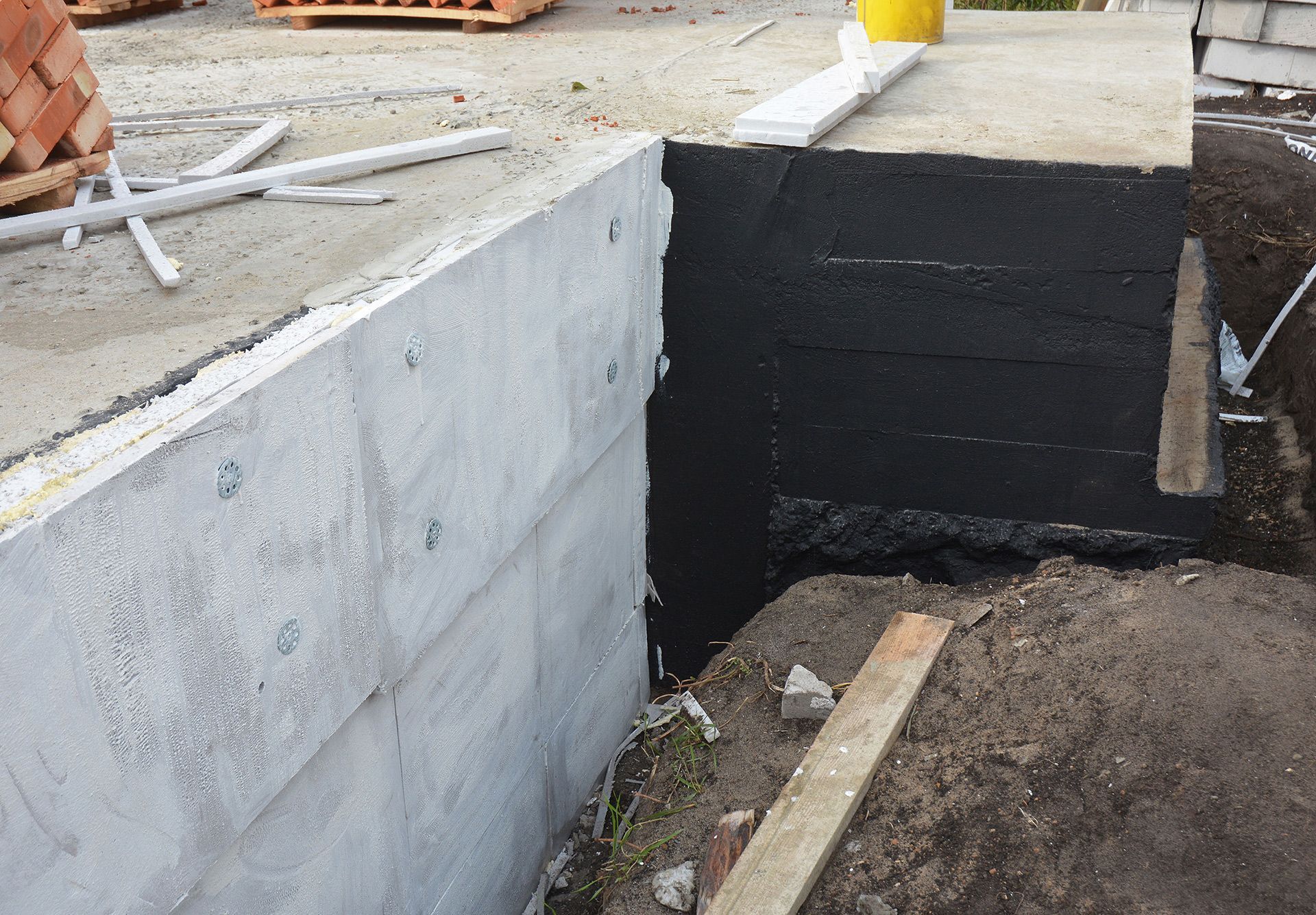 Concrete foundation wall with a black waterproofed corner beside a dirt trench at a construction site