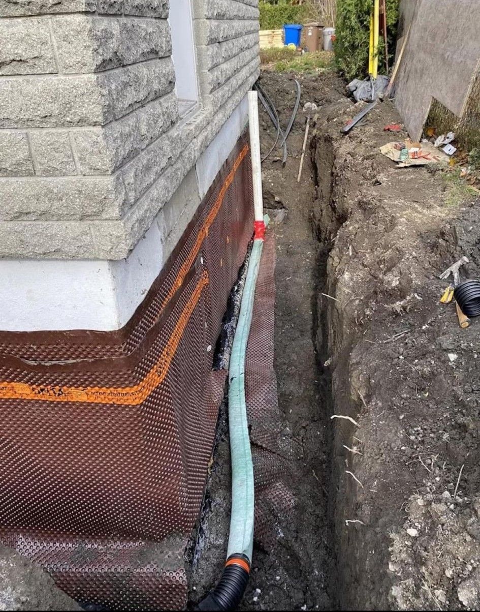 Foundation wall with orange waterproofing and a trench dug beside a stone building.