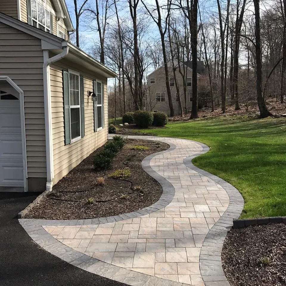 Curved stone walkway beside a house, bordered by mulch and lawn, leading through a wooded yard.
