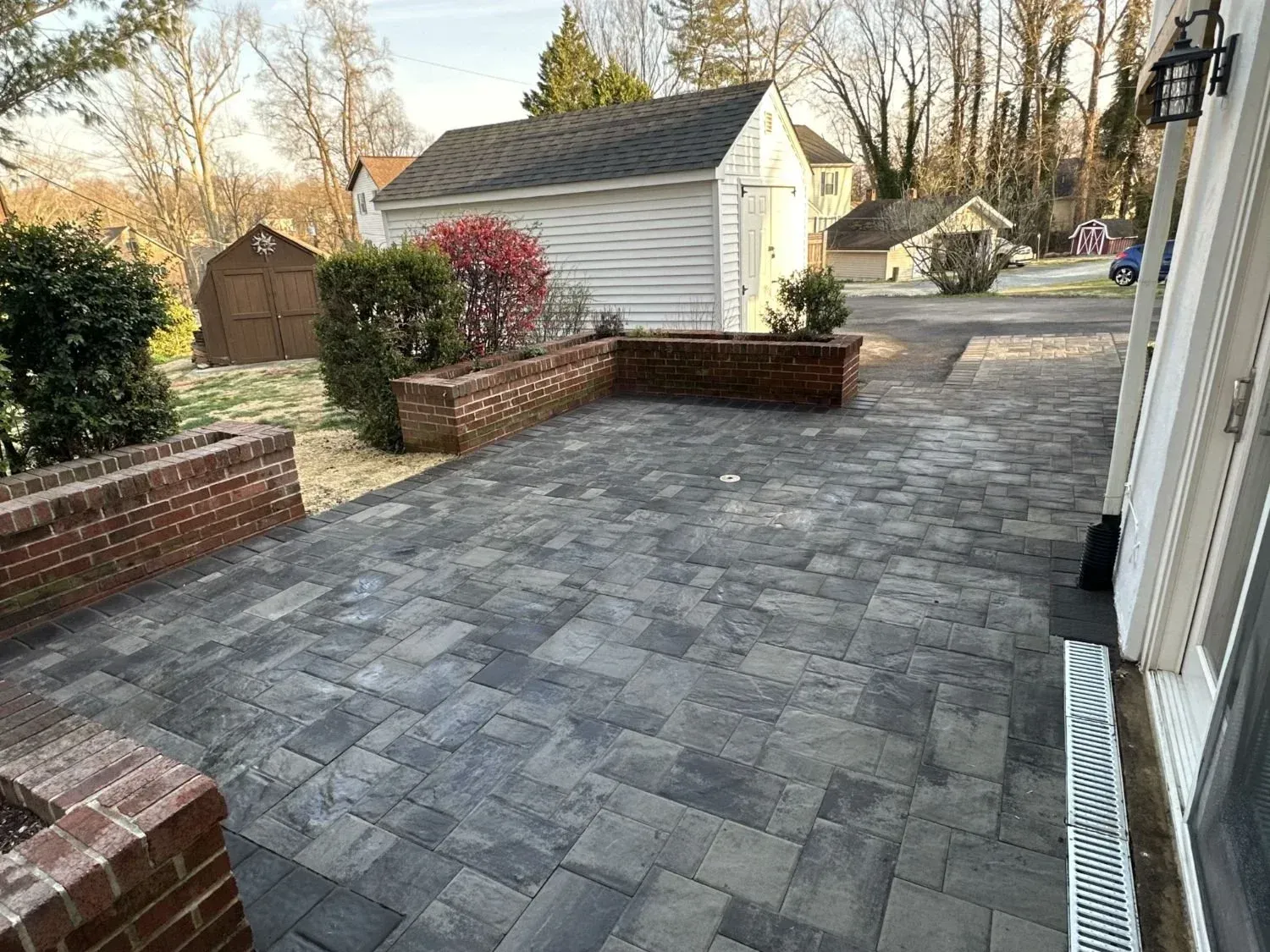 Stone-paver patio and driveway beside a white house, with low brick walls and a small shed in a backyard.