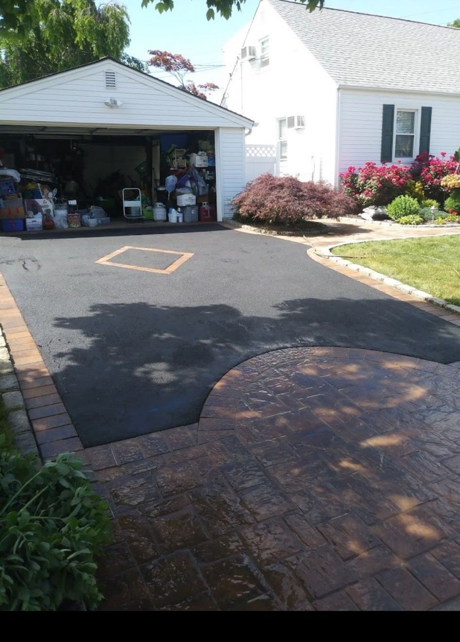 Driveway leading to a garage and white house, with a brick patio in the foreground and shrubs along the side.