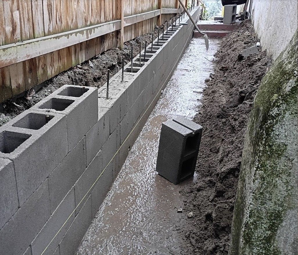 Narrow muddy alley with a retaining wall under construction and a loose concrete block in the foreground