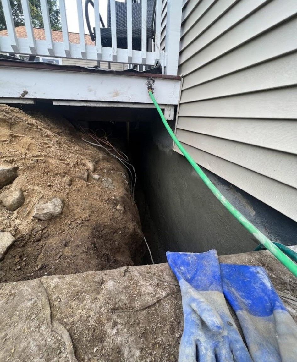 Green hose under a deck beside a house, with exposed soil and a blue tarp over a stone wall.
