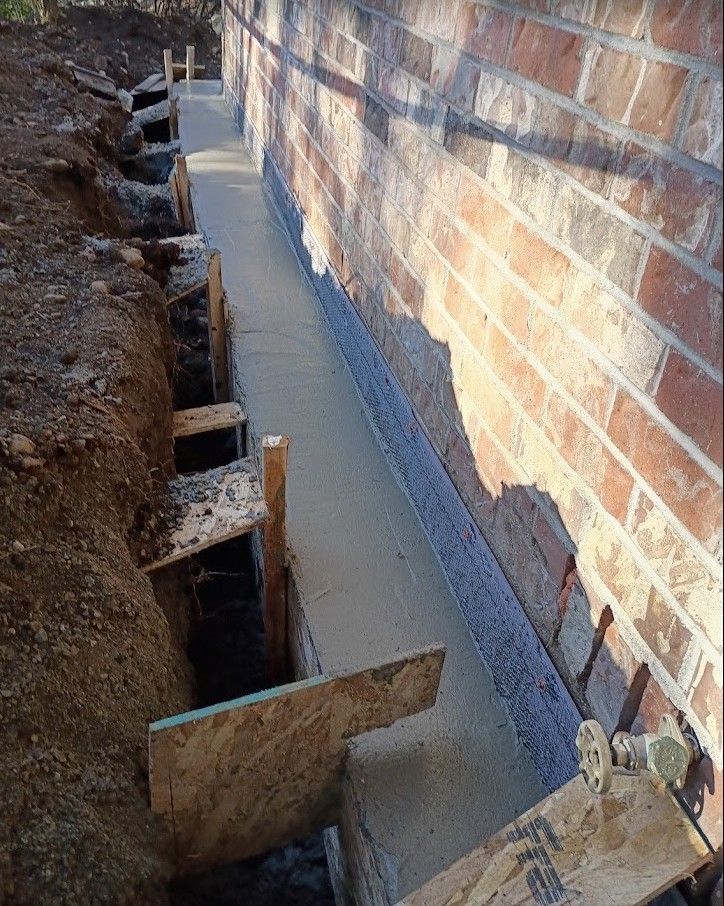 Fresh concrete footing poured beside a brick wall in an excavated trench with wooden braces.