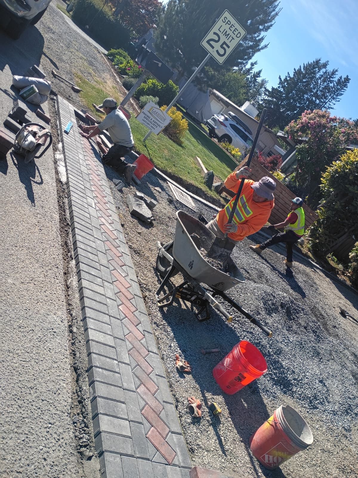 Road workers in orange vests edging a sidewalk beside a brick curb and gravel landscaping.