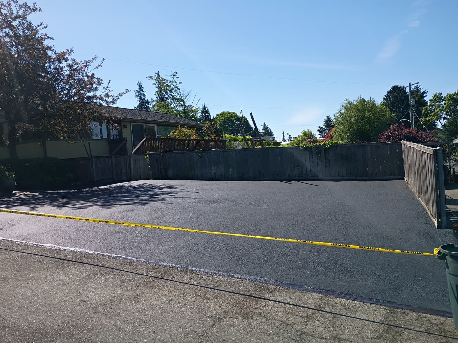 Empty asphalt parking lot with a yellow line, bordered by a wooden fence and trees under a clear sky