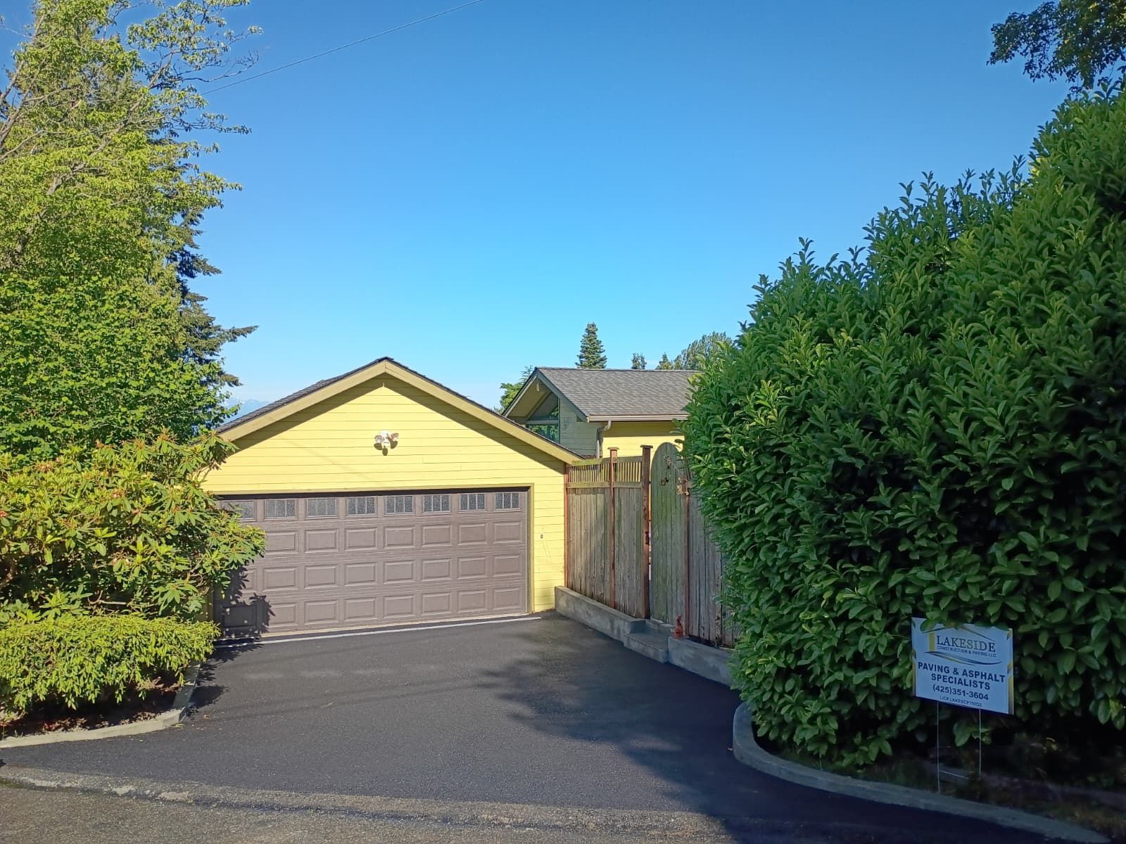 Suburban house with yellow garage, driveway, and tall hedges under a clear blue sky
