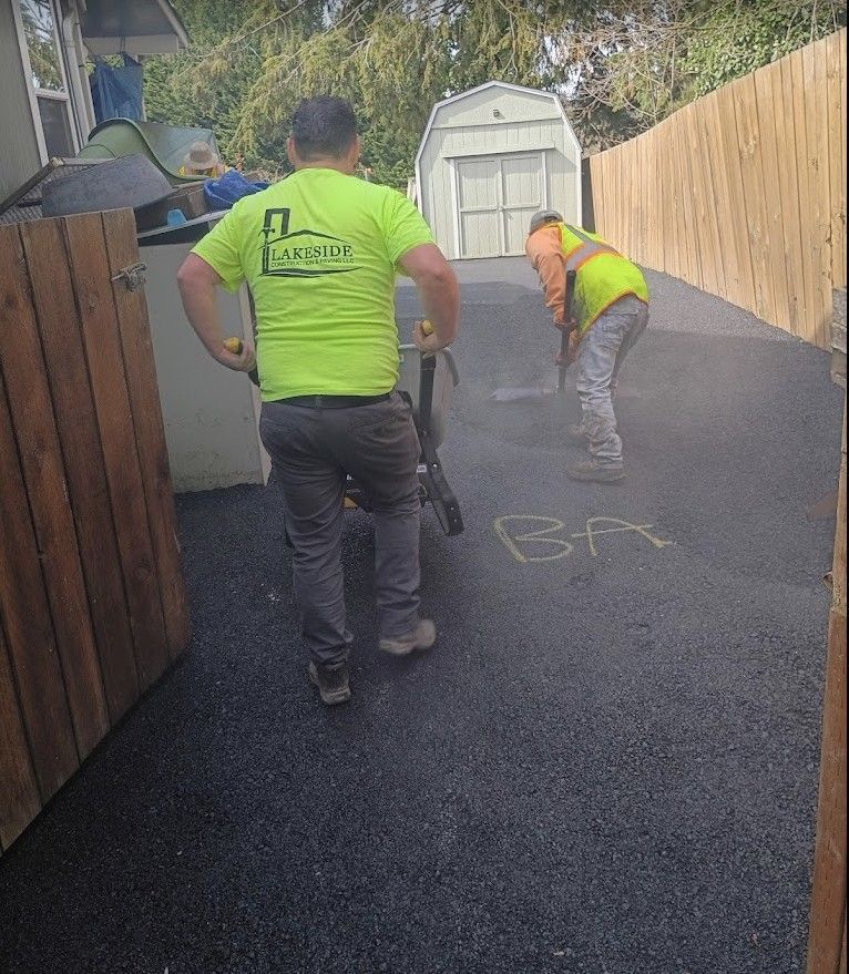 Two workers paving a narrow driveway beside a fence and shed, with one smoothing fresh asphalt.