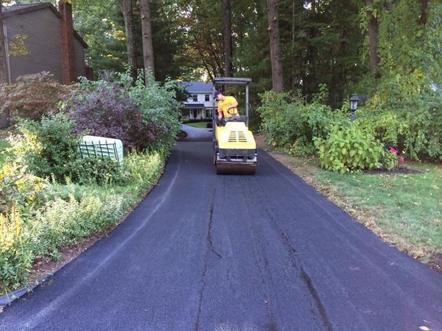 Person operating a small yellow road roller on a freshly paved driveway surrounded by trees and shrubs