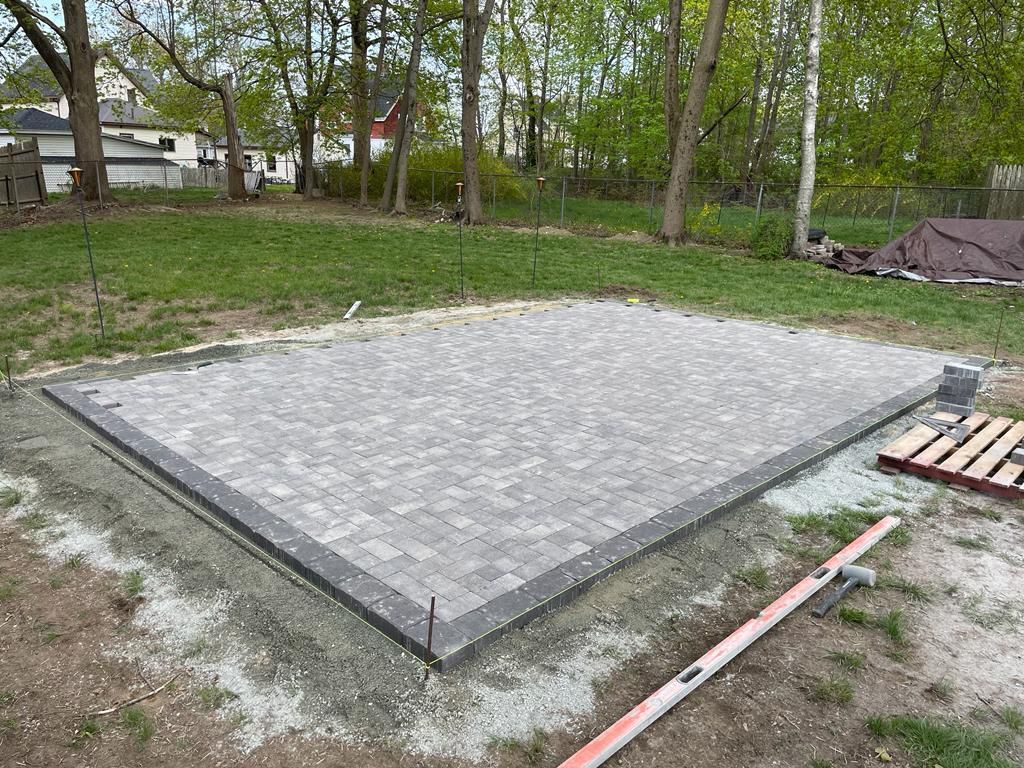 Newly paved rectangular gray patio foundation in a grassy backyard with trees and construction materials nearby