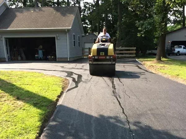 Person on a yellow lawn roller on a driveway beside a house, with a garage and trees nearby