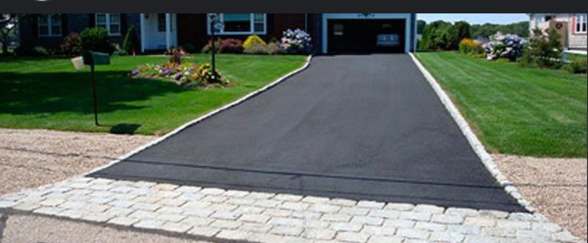 Black asphalt driveway leading to a house with a garage, bordered by stone pavers and green lawn