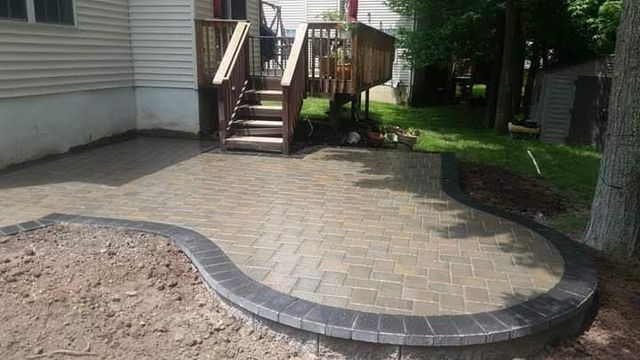 Curved stone paver walkway beside a house, leading to wooden steps and a shaded yard