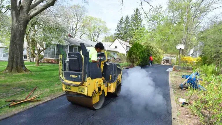 Yellow road roller paving a wet black driveway beside a suburban street, with steam or smoke rising