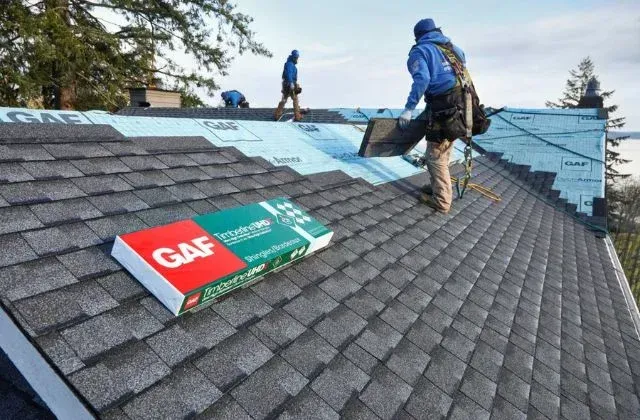 Roofers installing blue underlayment on a shingled roof, with a GAF roofing package in the foreground