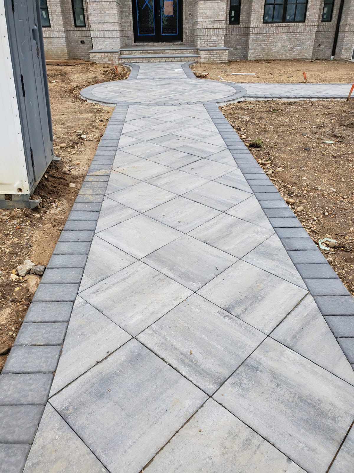 Diagonal concrete walkway leading to a house entrance, bordered by brick pavers and dirt landscaping.