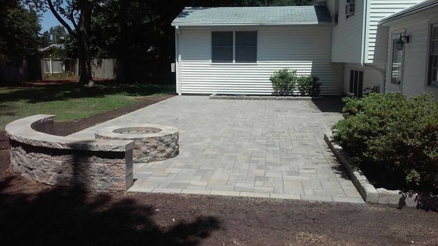 Stone paver driveway and patio beside a white house, with a curved retaining wall and shrubs