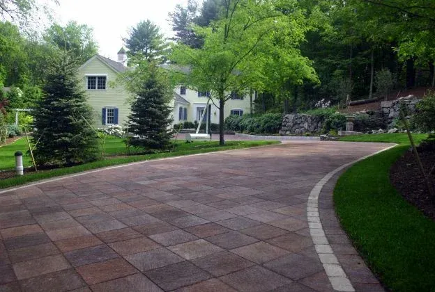 Curved brick driveway leading to a white house surrounded by green trees and shrubs