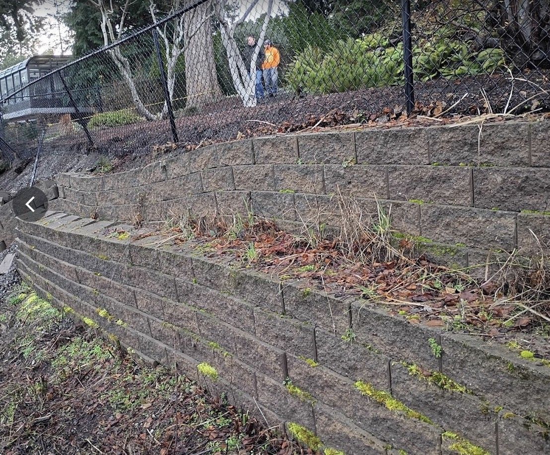 Terraced stone retaining wall beside a wooded path with a person in an orange jacket in the background