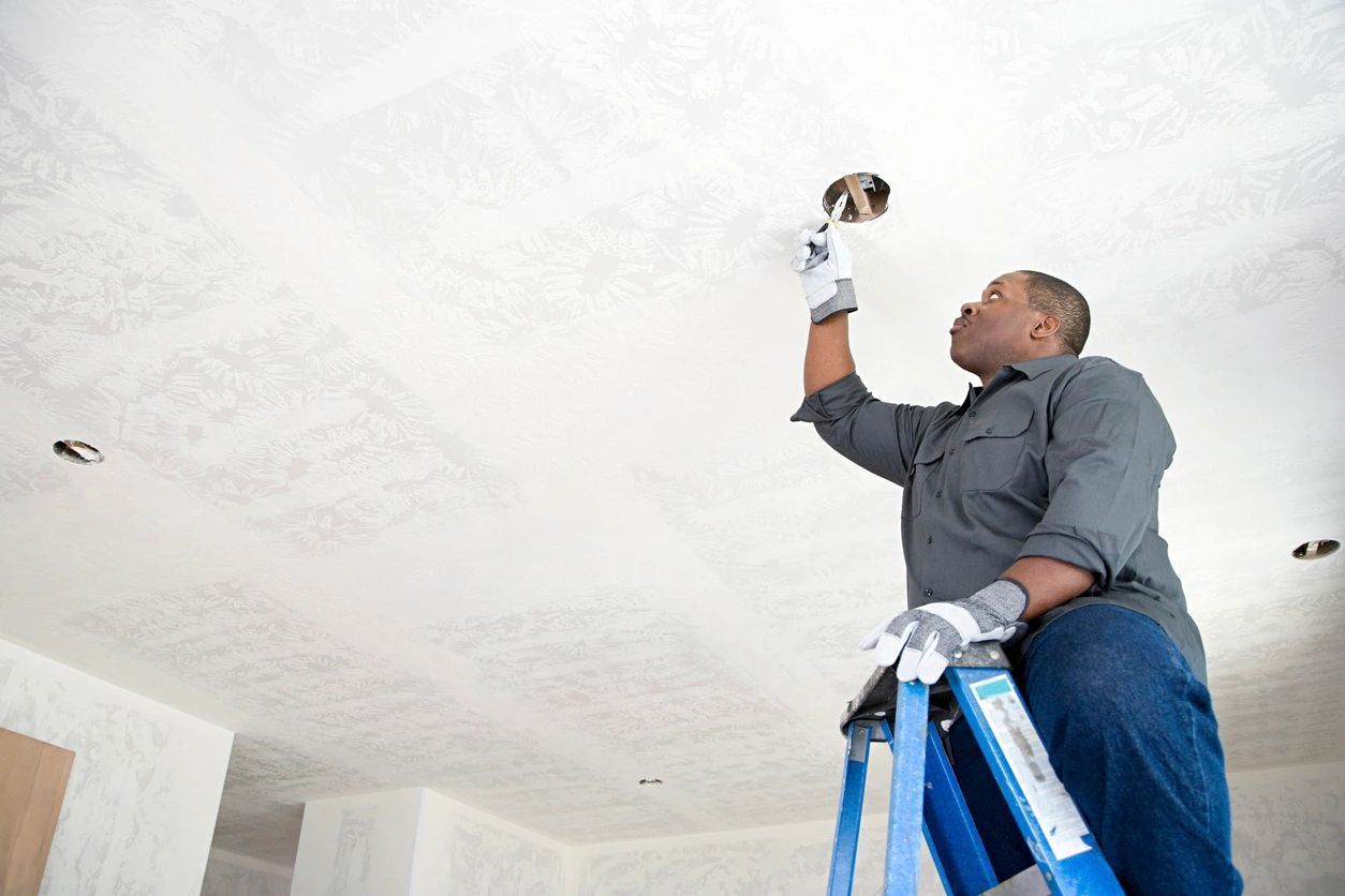 Person on a ladder working on a ceiling electrical fixture; wearing work gloves.