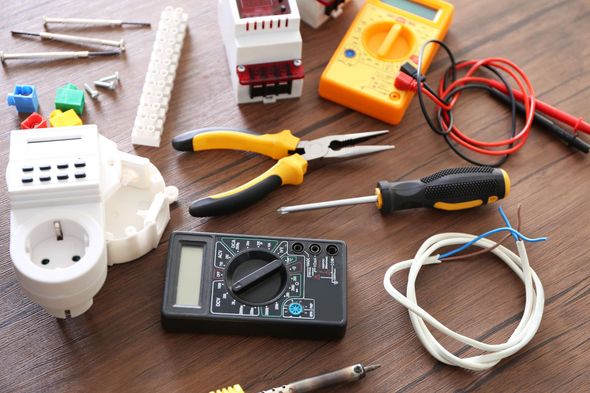 Electrical tools and components, including pliers, screwdriver, multimeter, and outlet, on a wooden surface.