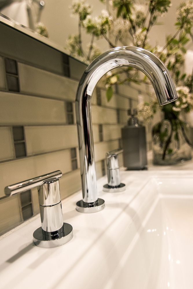 Chrome faucet on a white sink with a mosaic tile backsplash and flowers in the background.