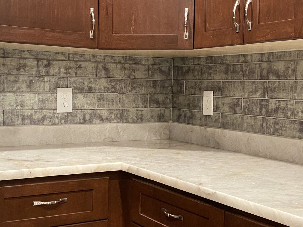 Brown kitchen cabinets, gray brick-like backsplash, and light countertops.