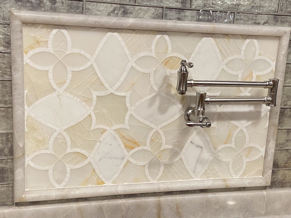 Decorative tile backsplash with a pot filler faucet. Off-white and beige floral pattern within a marble frame.