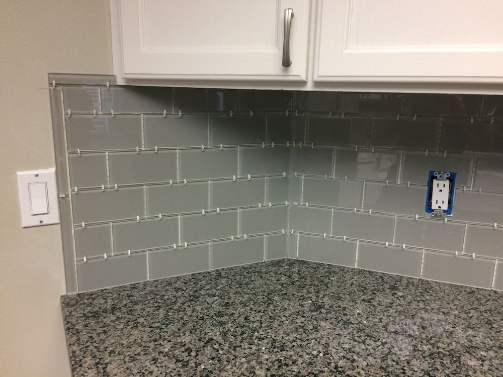 Gray tiled backsplash in a kitchen corner, with granite countertop and white cabinets.