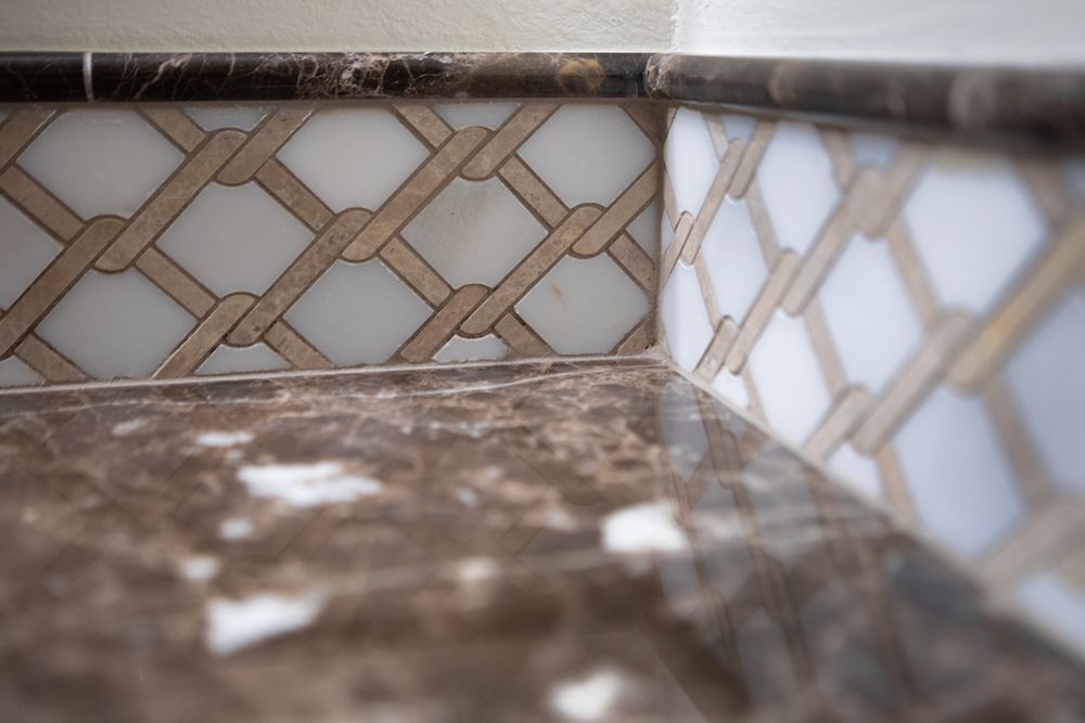 Close-up of a tiled countertop edge with a brown marble surface and woven mosaic pattern on the wall.
