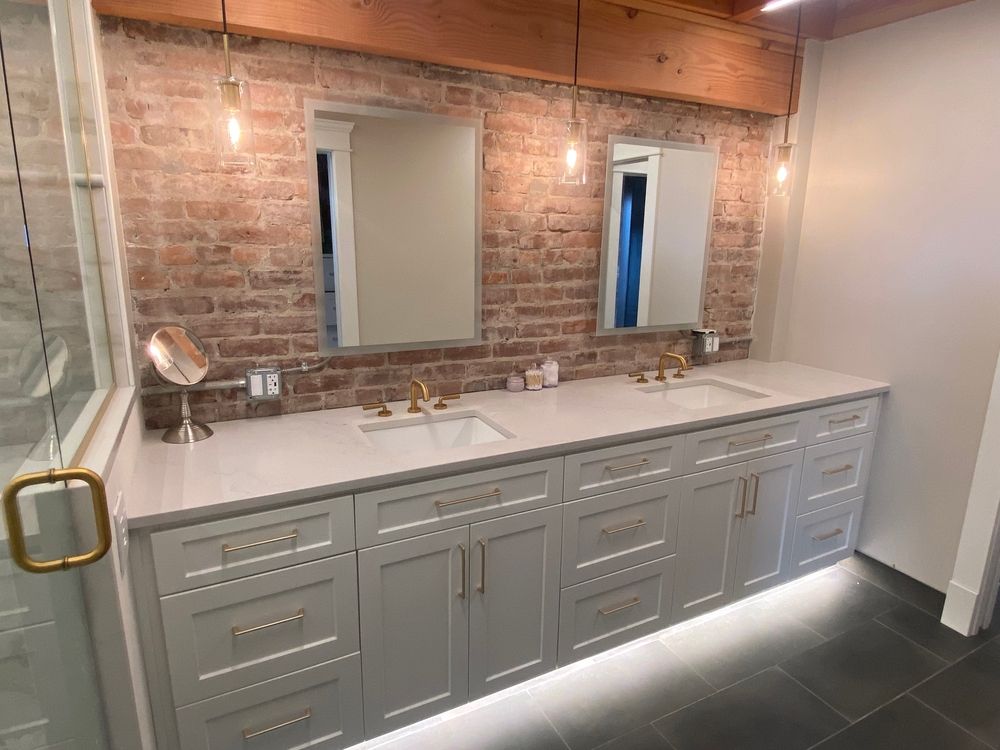 Bathroom with brick wall, double vanity, mirrors, gold hardware, and under-cabinet lighting.