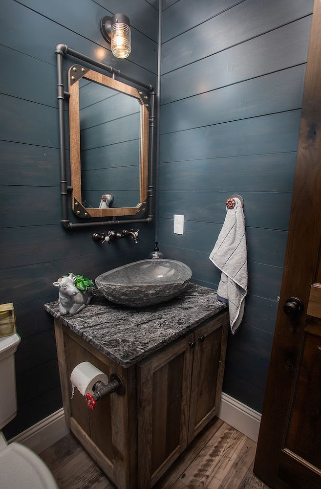 Rustic bathroom with wood vanity, stone sink, dark blue plank walls, and pipe-framed mirror.