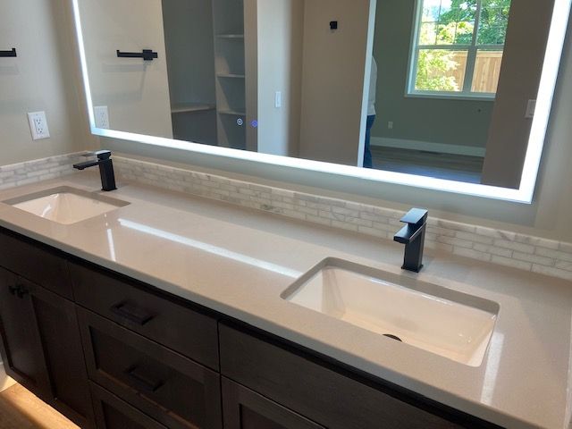 Bathroom with double vanity; dark brown cabinets, white countertop, black faucets, and large lighted mirror.