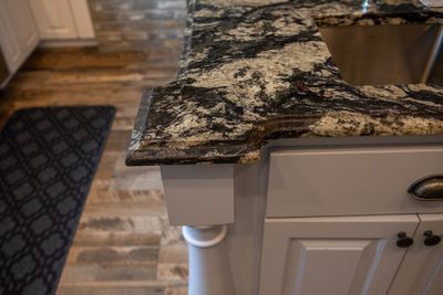 Close-up of a kitchen island with a granite countertop and white cabinets, near a wood floor and mat.