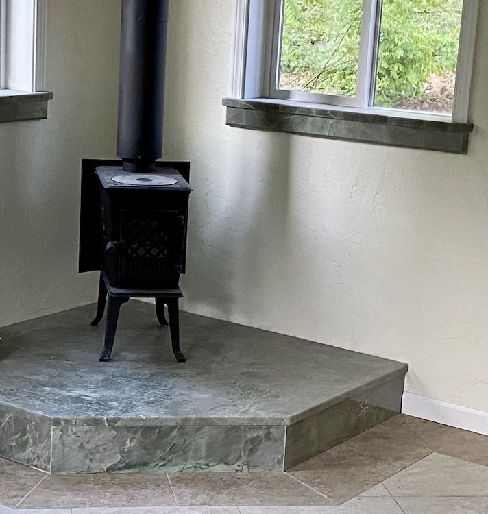 Black wood-burning stove on a raised tiled platform in the corner of a room, near a window.