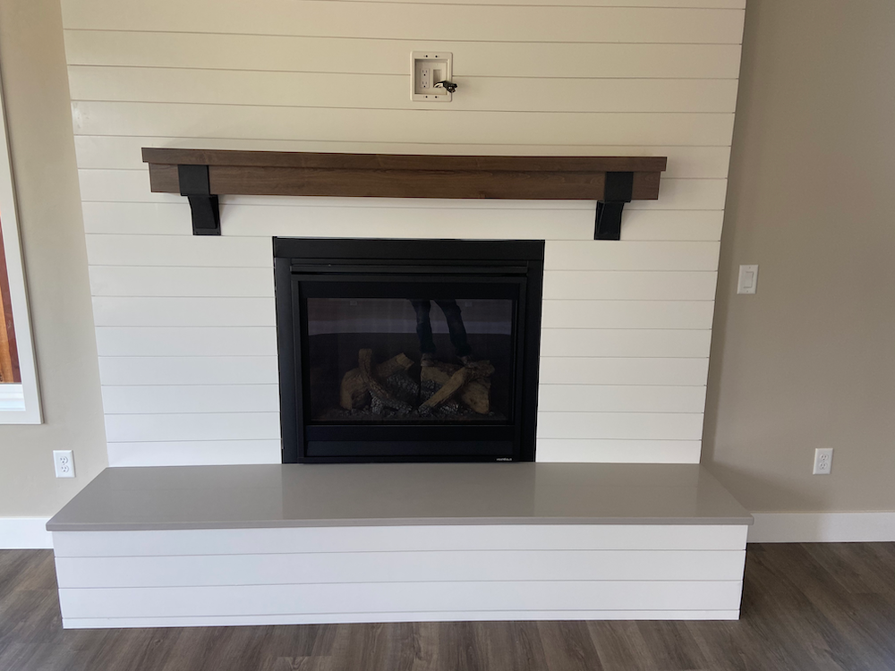 Fireplace with white shiplap, brown wooden mantel, black brackets, and a gray hearth in a living room.